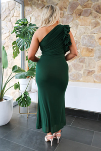 back view of a woman in a green one-shoulder dress standing in a room with stone walls and plants.