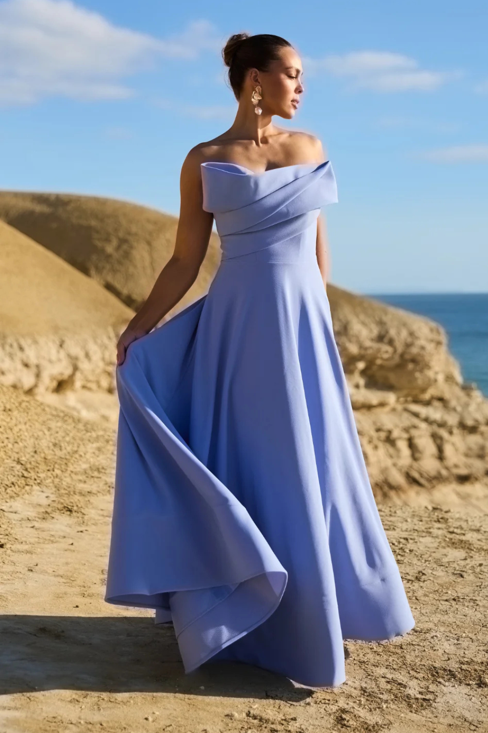 Woman in a blue off-shoulder dress standing on a beach with ocean and sky in the background
