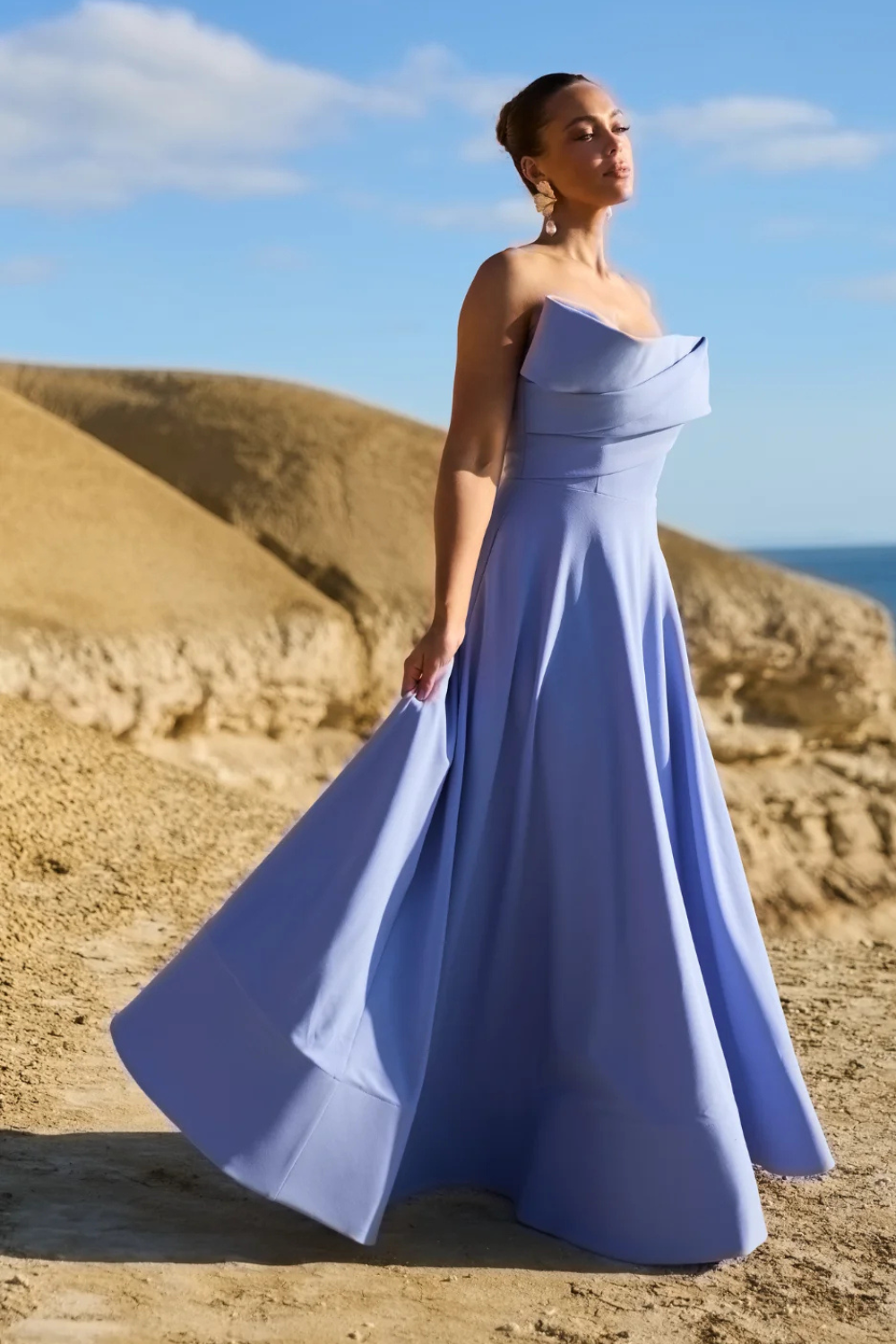 Woman in a blue gown standing on a desert landscape with hills and clear sky.