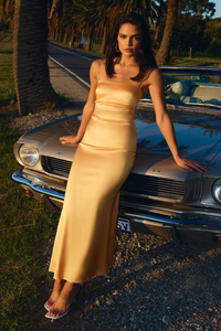 A model in a low-cut, sleeveless, yellow dress with a straight neckline and adjustable straps, standing next to a vintage car.