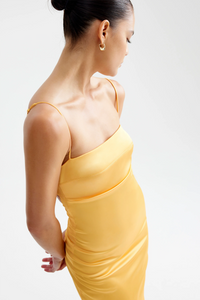 Close-up profile view of a model wearing the yellow Studio LEXI Bora Dress, showcasing the straight neckline, delicate spaghetti straps, and gold hoop earring against a white studio background.